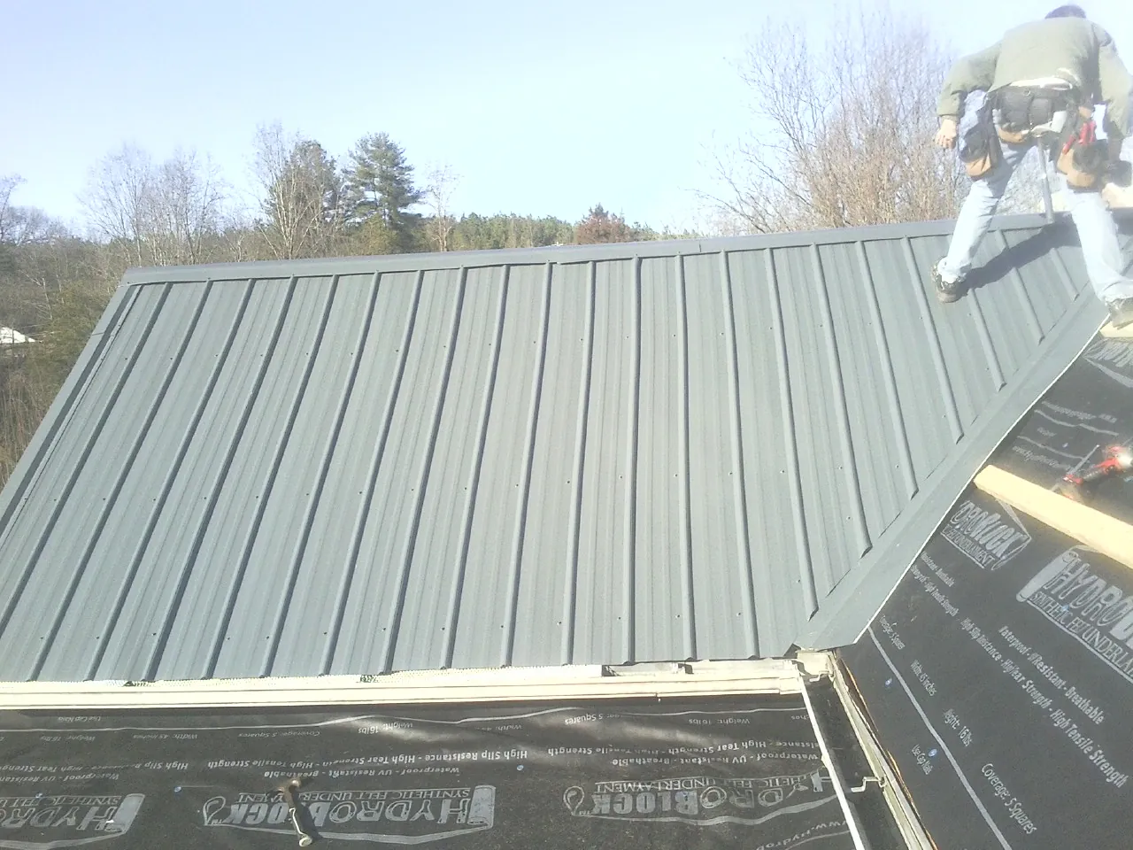 Crew installing dark green standing seam panels over synthetic underlayment in Ulmer