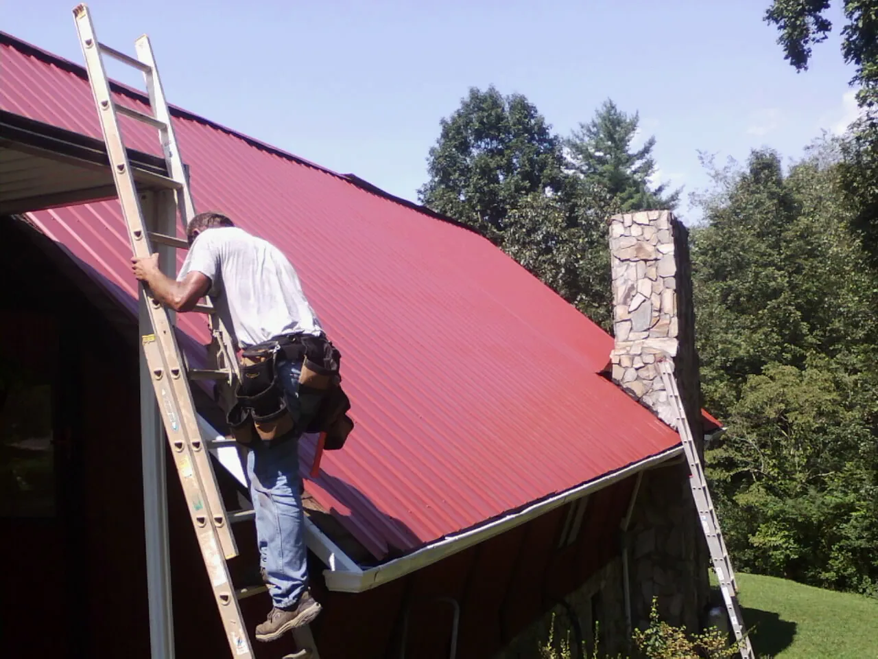 Roofer on a ladder beside a red metal roof and stone chimney — Soffit & Fascia Repair by Roofing Brothers in Ulmer